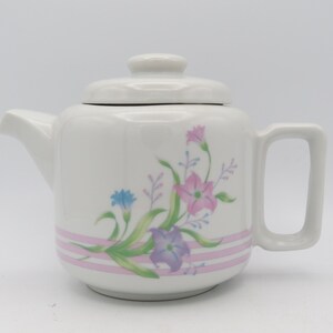 May include: A white ceramic teapot with a floral design featuring pink, purple, and blue flowers. The teapot has a lid and a handle.