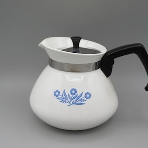 May include: White coffee pot with a black handle and lid. The pot has a silver band and a blue floral design. The spout is white and the handle is curved. The pot is likely made of ceramic.