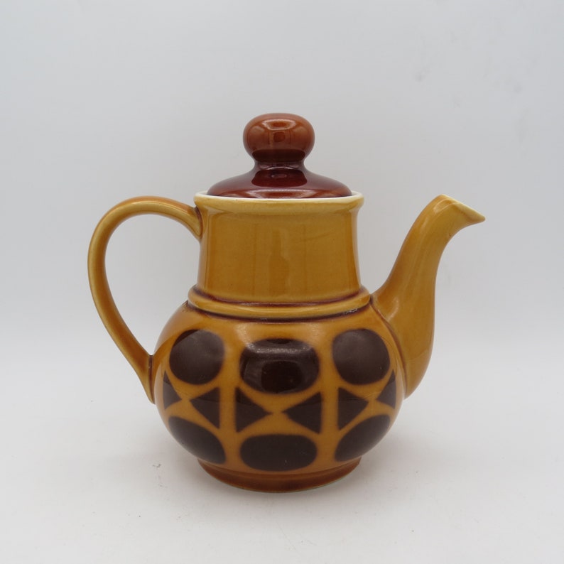 May include: A brown and yellow ceramic teapot with a geometric pattern of brown circles and triangles. The teapot has a lid with a brown knob.