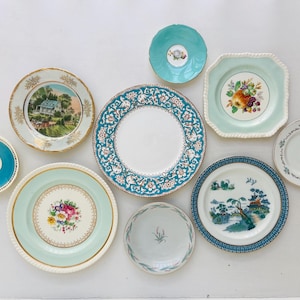 May include: A collection of nine ceramic plates and saucers in various sizes, featuring diverse designs including floral patterns, landscape illustrations, and solid teal or mint green borders with gold trim accents arranged on a flat surface.