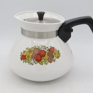 May include: White enamel teapot with a black handle and lid. The teapot features a decorative floral design with yellow, brown, and red fruits and flowers. A silver band encircles the middle of the teapot.