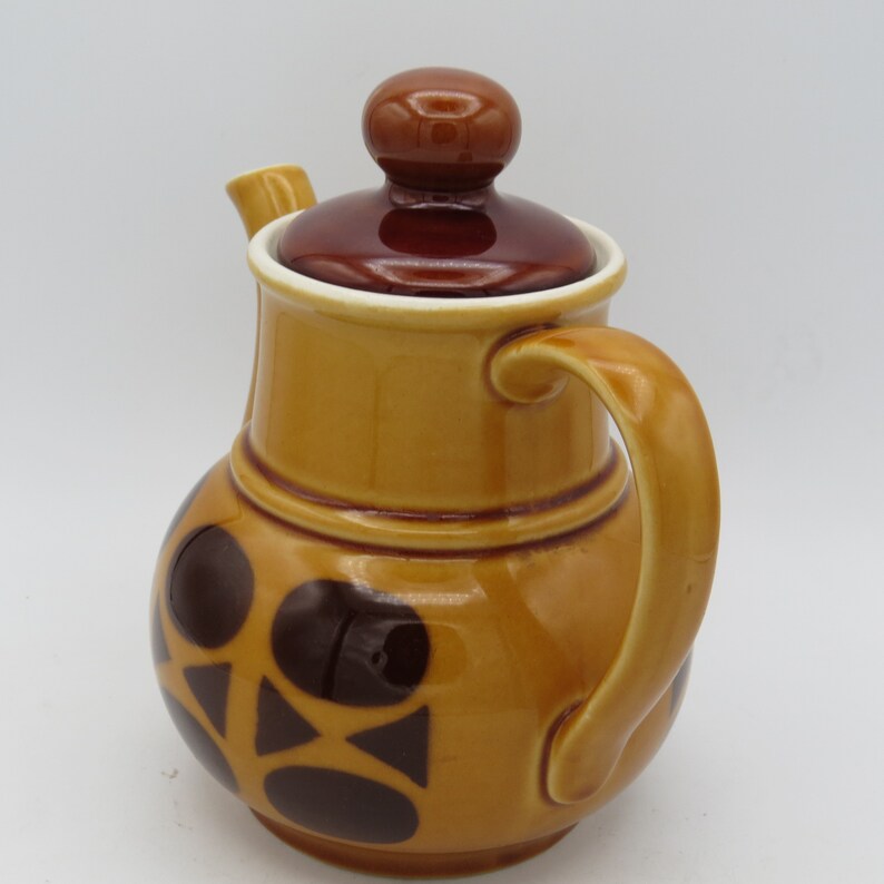 May include: A brown ceramic teapot with a brown lid and a brown handle. The teapot has a geometric pattern of brown circles and triangles on a light brown background.