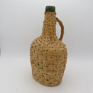 May include: A glass bottle with a dark green neck and opening, fully encased in a light tan woven wicker covering. The exterior features a braided wicker handle attached to the neck and the main body, providing a textured, basket-like appearance.