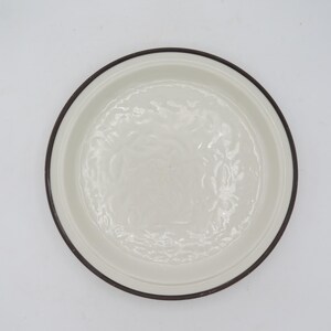 May include: A white ceramic plate with a dark brown rim. The plate has a slightly raised edge and a floral pattern embossed in the centre. The plate is round and appears to be new.