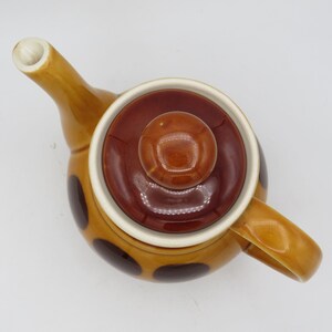 May include: A brown and yellow teapot with a brown lid and a brown knob. The teapot has a polka dot pattern in brown.