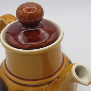 May include: A brown and tan ceramic teapot with a brown lid and a brown handle. The teapot has a brown and white pattern on the body.