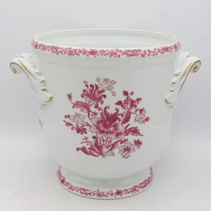 Könnte beinhalten: Weißer Keramik-Blumentopf mit einem floralen Design in Rosatönen. Der Pflanztopf hat zwei verzierte Griffe mit goldenen Akzenten und einen dekorativen rosa Rand um den Rand und die Basis. Ein klassisches Stück für die Inneneinrichtung.