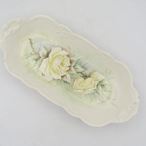 Könnte beinhalten: Eine weiße Keramik-Servierplatte mit gewelltem Rand. Die Platte ist mit einem handgemalten Design aus zwei gelben Rosen mit grünen Blättern verziert.