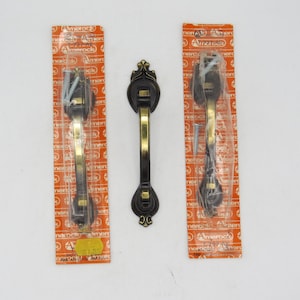 May include: Three black and gold cabinet door pulls with ornate designs. Each pull comes with two screws packaged on a card with the brand name "Amerock" printed on it.