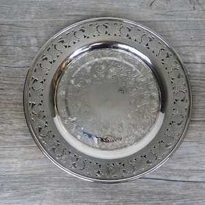 May include: A silver plate with an intricate design. The plate has a detailed floral and vine pattern around the edge, with an embossed design in the centre. The plate is circular and appears to be made of metal.