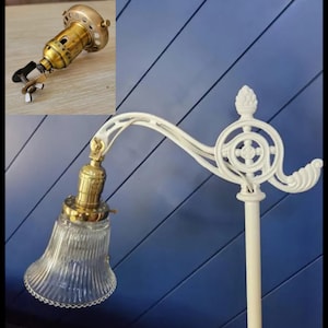 May include: A white metal lamp arm with a decorative finial and a brass lamp socket holding a clear, ribbed glass shade. An inset shows a close-up of the brass lamp socket with a metal clamp and wing nut attachment for mounting the fixture.