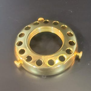 May include: A circular, polished gold-tone metal lamp shade holder featuring a central threaded opening surrounded by a ring of evenly spaced circular cutouts. Three small, adjustable metal screws are positioned around the outer edge.