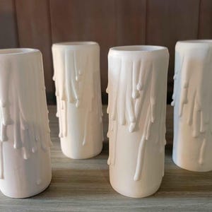 May include: Four off-white candle holders with a dripped wax design. The cylindrical holders are arranged on a wooden surface. The wax appears to be dripping down the sides of the holders, creating a textured effect.