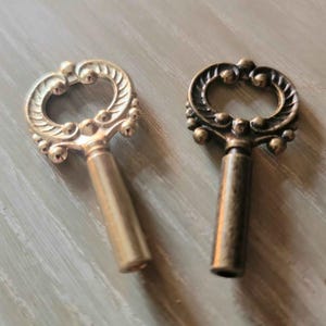 May include: Two decorative antique-style keys. One key is gold-colored, the other is bronze. Both keys have ornate, openwork tops with a loop design and small ball accents. The keys are on a gray surface.