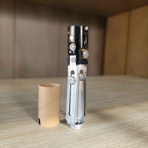 May include: A disassembled black and silver metal object with screws and a small brown cardboard tube. The object has a complex design with multiple parts and appears to be a component of a larger device.