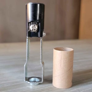 May include: A black lamp socket with a silver metal bracket and a small cardboard tube. The lamp socket has a screw and a silver metal base. The cardboard tube is brown and cylindrical.