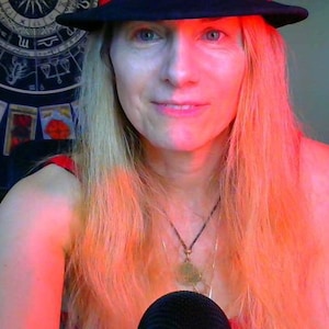 May include: A woman with long blonde hair wearing a black fedora and a red tank top. She is wearing a necklace with a pendant. She is looking at the camera and smiling.