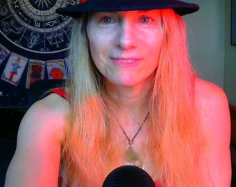 May include: A woman with long blonde hair wearing a black fedora and a red tank top. She is wearing a necklace with a pendant. She is looking at the camera and smiling.