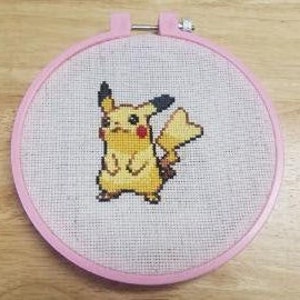 May include: A pink embroidery hoop with a cross-stitch design of Pikachu, a yellow electric-type Pokemon with black ears and a tail.