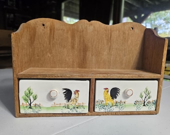 Puede incluir: Un estante de madera para pared con dos cajones de cerámica blanca. Cada frente de cajón está decorado con una escena pintada a mano de un gallo, un árbol pequeño y una valla. La unidad tiene un borde superior festoneado y dos orificios de montaje.
