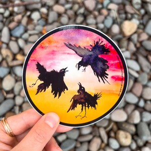 May include: A round sticker with a watercolor background of pink, orange, and yellow. Three black crows are silhouetted against the sunset. The sticker has the text "erin lacera art | erinlacera.com | @erinlaceraart" on the right side.