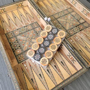 Può includere: Un tavolo da backgammon in legno aperto con motivi decorativi intarsiati. Il tavolo è realizzato in legno chiaro e scuro, con intricati disegni geometrici. Sono visibili pedine e dadi.