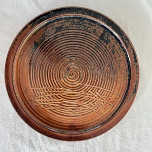 May include: A circular ceramic plate featuring a spiral pattern at its core. The plate is predominantly brown and black, with a textured finish. It has a raised edge and an etched decorative element.