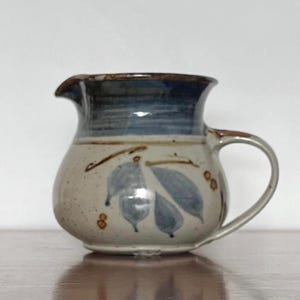 May include: A ceramic jug with a rounded body and a handle. The jug is beige with blue and brown accents, including a blue band around the rim and blue leaf designs. It features a spout for pouring liquids.