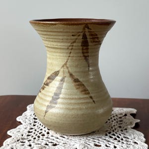 May include: A beige ceramic vase with a brown rim and a leaf design. The vase has a textured surface with horizontal lines and a wide opening. The leaf design is in shades of brown.