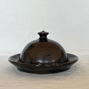 May include: A dark brown ceramic butter dish with a domed lid and a round base. The lid has a small knob on top. The dish is designed to hold butter and keep it fresh. The glossy finish gives it a classic look.