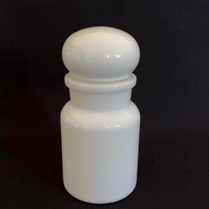 May include: A white apothecary jar with a rounded lid. The jar has a cylindrical body and a slightly flared base. The lid is a smooth, spherical shape. The jar is made of opaque white glass or ceramic.