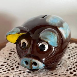 May include: A ceramic piggy bank shaped like a pig. The bank is brown with blue and yellow accents on the ears and snout. The eyes are white with black pupils. There is a coin slot on the top.