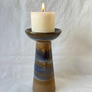 May include: A burning candle rests on a ceramic candle holder. The holder has a brown base, a blue and brown glazed body, and a dark brown rim. The candle is ivory-coloured and cylindrical, with a visible flame.