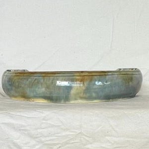 May include: A ceramic bowl with an oval shape. The bowl has a mottled glaze in shades of blue, green, and brown. Small, raised handles are on each end of the rim. The bowl is likely handmade and suitable for serving or display.