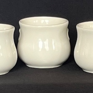 May include: Three white ceramic cups with a rounded, bulbous shape. Each cup has a small, raised design on the side.