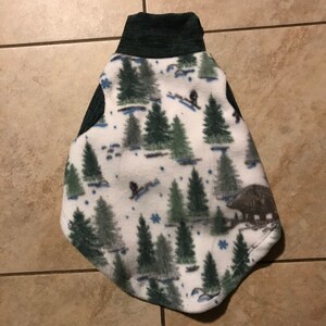 May include: White fleece dog vest with a green and blue winter forest print. The vest has a dark gray ribbed collar.