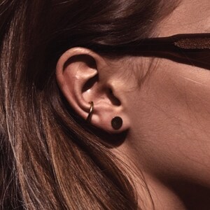 May include: A woman with long brown hair wearing black sunglasses and small gold hoop earrings. She is wearing a black shirt and has a dark brown stud earring in her left ear.