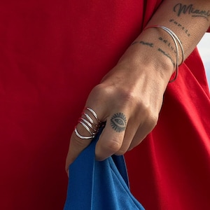 Close-up of a hand pulling up a blue fabric, showcasing silver spiral rings against a vibrant red background, complementing a small eye tattoo on the wearer's finger.