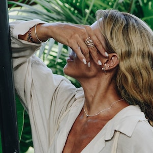 A woman in a relaxed pose with her head tilted back and hand on forehead, showcasing multiple silver rings and ear cuffs, set against a lush green background.