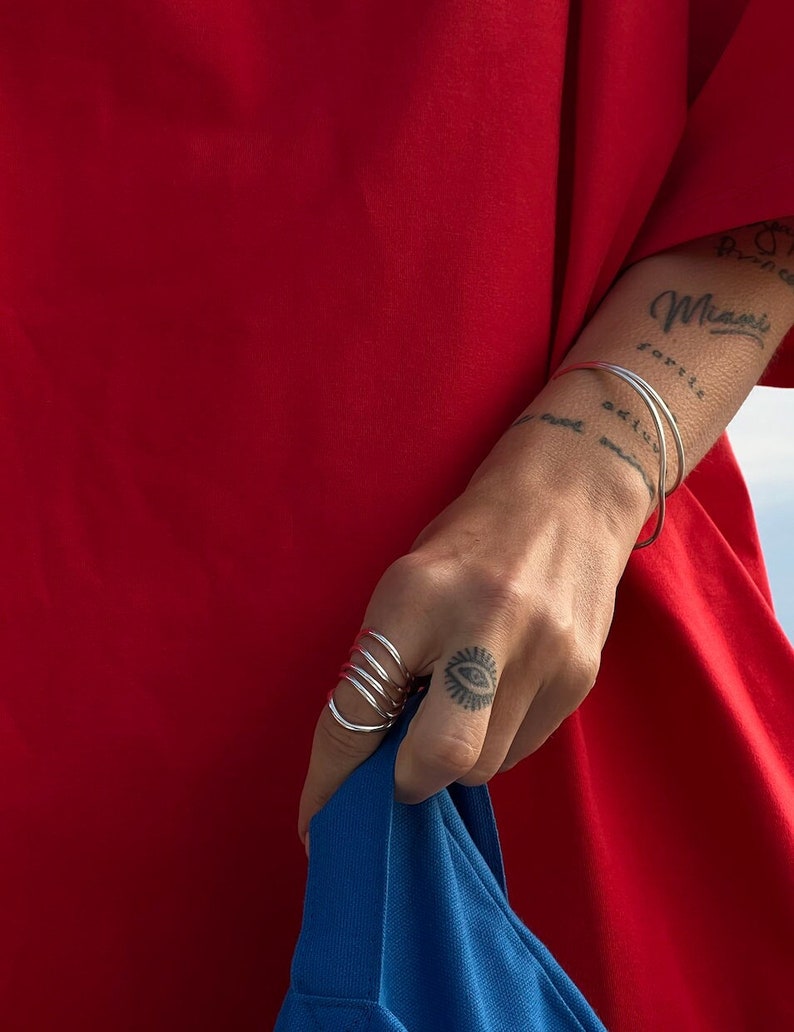 Close-up of a hand pulling up a blue fabric, showcasing silver spiral rings against a vibrant red background, complementing a small eye tattoo on the wearer's finger.