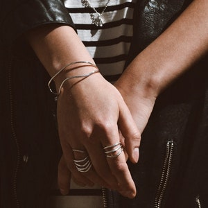 A hand with a silver bracelet and rings, including a prominent spiral ring, resting casually against a black leather jacket with striped clothing underneath.