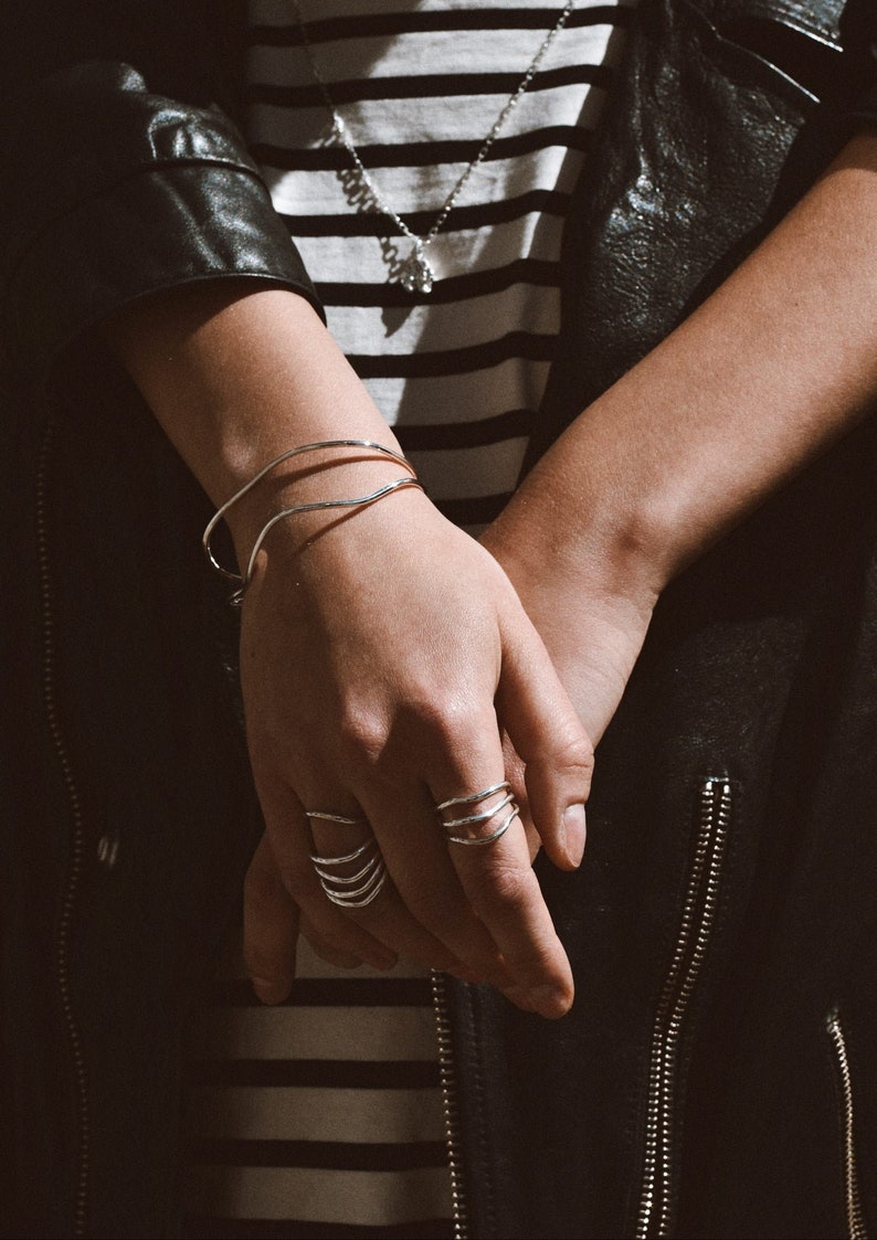 A hand with a silver bracelet and rings, including a prominent spiral ring, resting casually against a black leather jacket with striped clothing underneath.