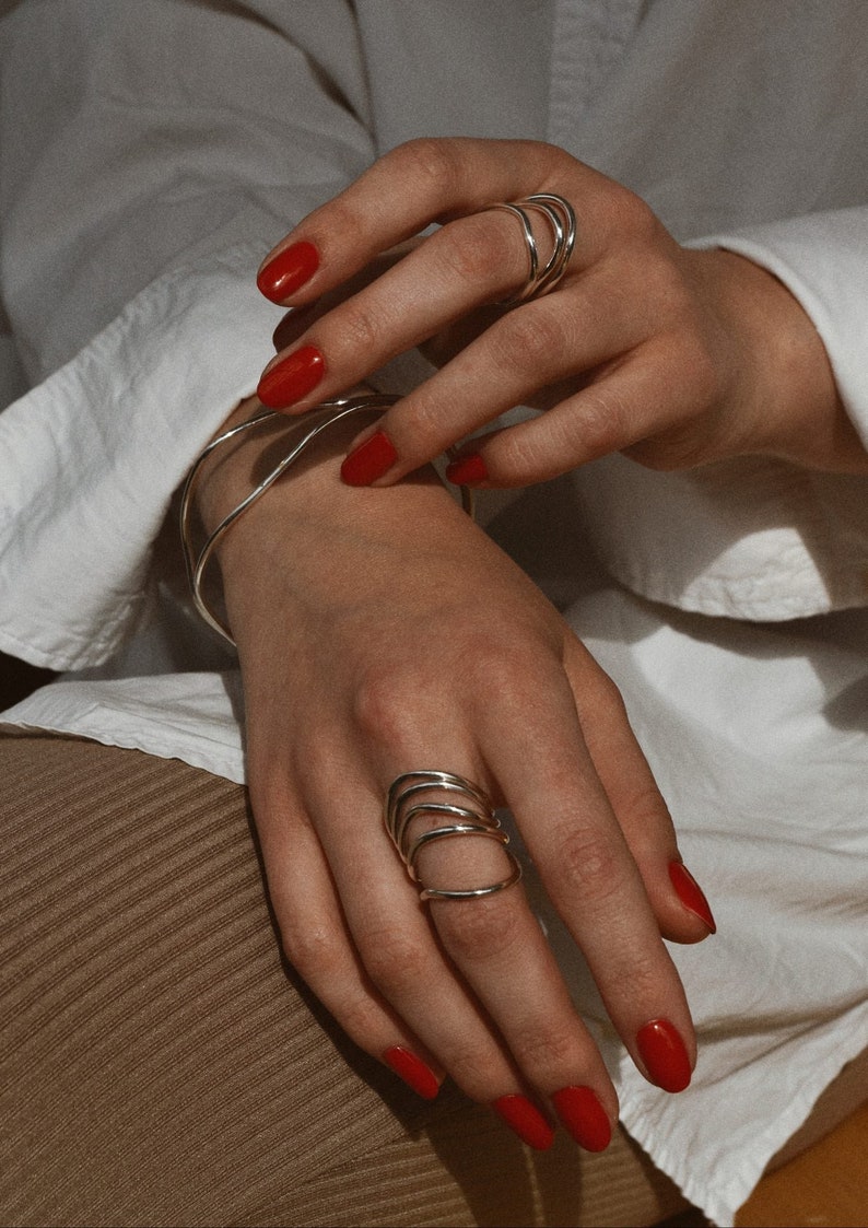 A hand with red nail polish displaying silver spiral rings, leaning against a knee covered in a white cloth