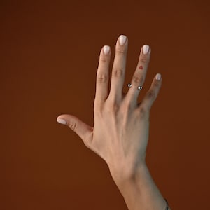 May include: A hand with a silver ring on the ring finger and a silver bracelet on the wrist. The hand is raised with the fingers spread apart against a brown background.