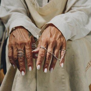 Artistic photo of hands clad in oversized linen sleeves, adorned with various silver rings and a connected chain ring, against a backdrop of soft, natural fabric