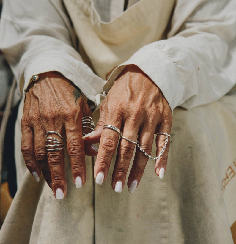 Artistic photo of hands clad in oversized linen sleeves, adorned with various silver rings and a connected chain ring, against a backdrop of soft, natural fabric