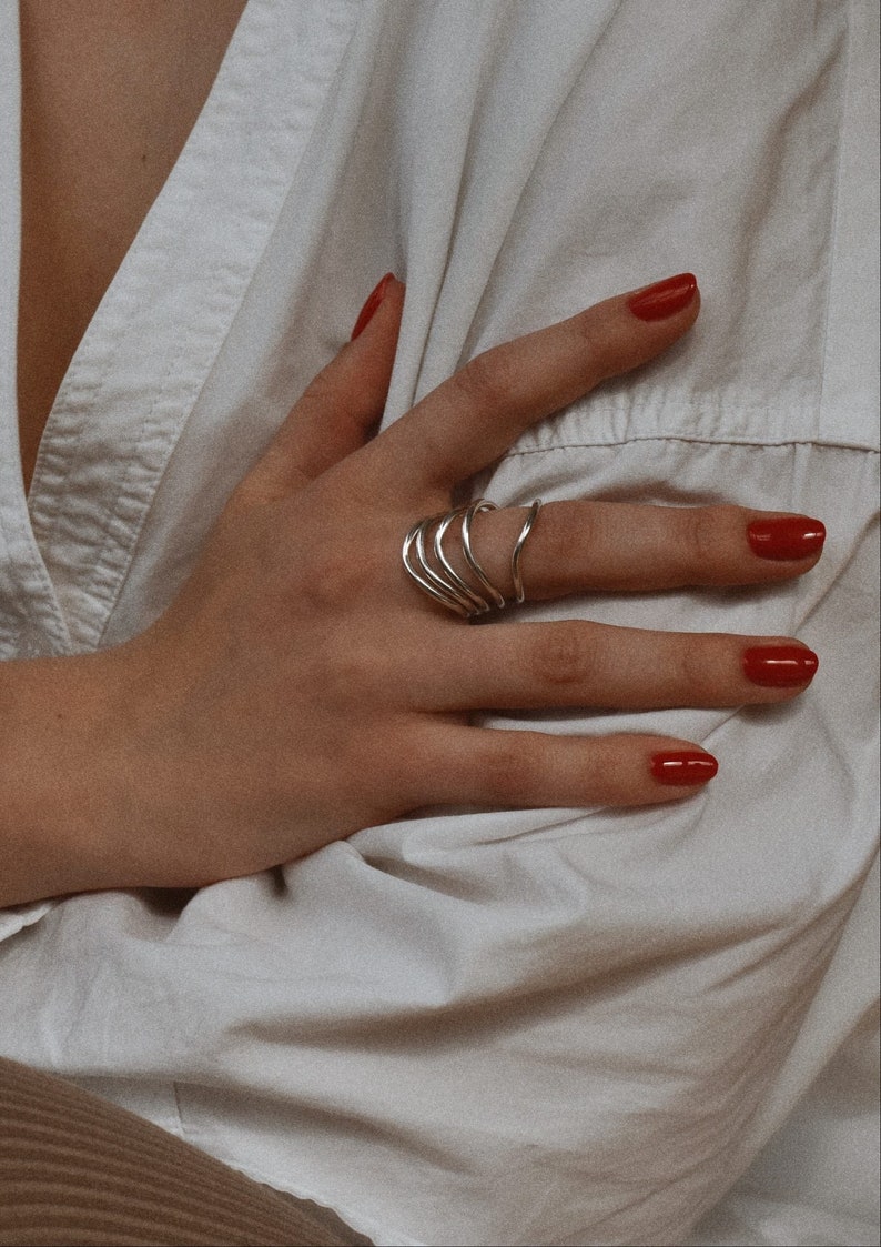 A close-up of a hand with red nail polish on a white cloth background, showcasing layered silver spiral rings.