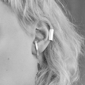 May include: A close-up of a person's ear with two silver ear cuffs. The ear cuff on the top is a simple, straight bar, while the ear cuff on the bottom is a curved hoop.