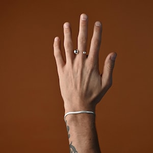 May include: A silver ring with two round balls on the top. The ring is worn on the middle finger of a hand with a silver bracelet on the wrist.
