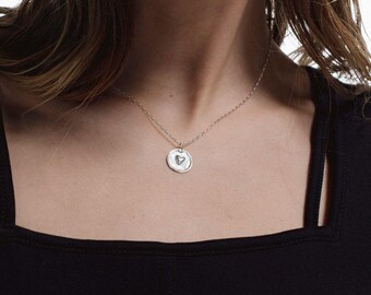 May include: A silver necklace with a small, round pendant featuring a heart design. The pendant hangs from a delicate chain. The person is wearing a black top.